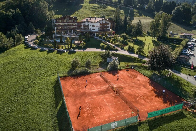 Tennisplatz Bichlhof Luftaufnahme von zwei Sandtennisplätzen vor einem Hotelgebäude in Hanglage.
