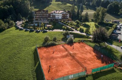Tennisplatz Bichlhof Luftaufnahme von zwei Sandtennisplätzen vor einem Hotelgebäude in Hanglage.