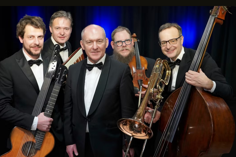 Fünf Musiker in schwarzen Anzügen mit Kontrabass, Posaune, Geige, Kontragitarre und Akkordeon vor blauem Hintergrund.