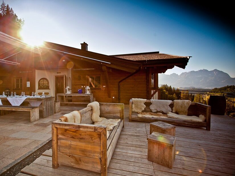 Holzterrasse einer Almhütte mit Sitzmöbeln aus Holz und Blick auf die Berge bei Sonnenuntergang.