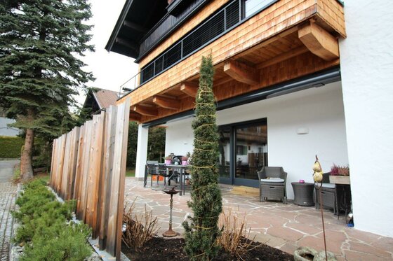 Gepflasterte Terrasse mit Gartenmöbeln vor einem modernen Haus mit Holzfassade und Balkon.