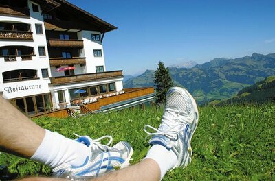 Beine in Sportschuhen auf einer Wiese vor einem Berghotel mit Blick auf die Alpen.