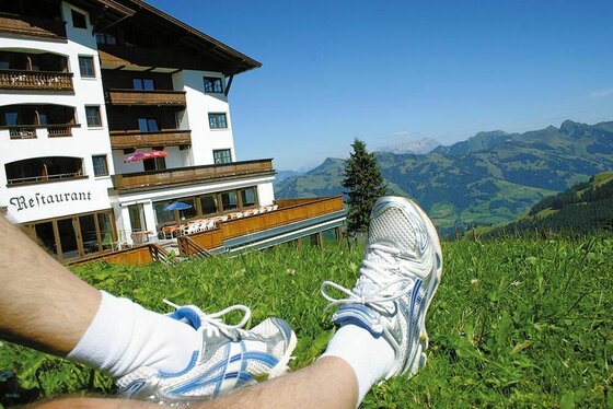 Sommer am Hotel Ehrenbachhöhe Beine in Sportschuhen auf einer Wiese vor einem Berghotel mit Blick auf die Alpen.