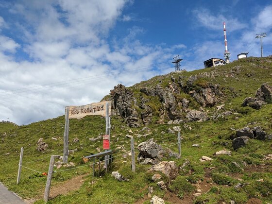 Kids Kletterpfad am Kitzbüheler Horn Holzportal mit der Aufschrift Kids Kletterpfad auf einer grünen Bergwiese vor felsigem Hang und Sendemast.