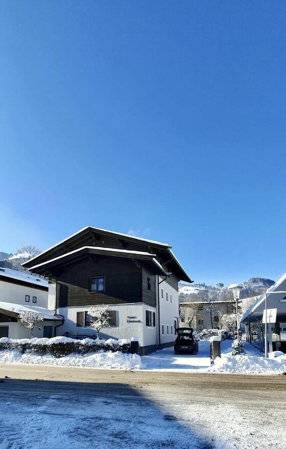 Zweistöckiges Haus mit dunkler Holzfassade im Obergeschoss in einer verschneiten Winterlandschaft unter blauem Himmel.