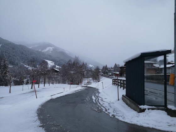 Verschneite Straße mit Bushaltestelle und bewaldeten Bergen im Hintergrund bei bewölktem Himmel.