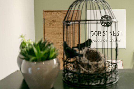 Eingangsbereich Doris' Nest Dekorativer Vogelkäfig mit Nest im Vordergrund, dahinter eine Holztür und ein Logo an der Wand.