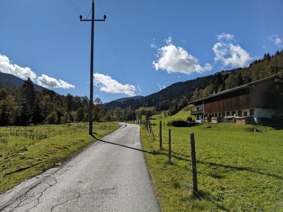 Jochberger 7-Brunnenweg Asphaltierter Weg führt an einem Bauernhaus und Strommasten vorbei durch eine Berglandschaft.
