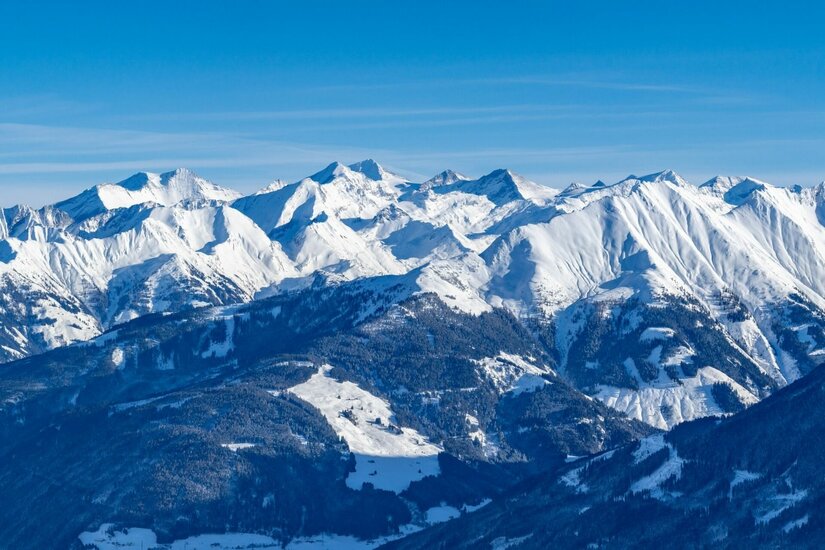 Verschneite Bergkette mit bewaldeten Hängen und Skipisten unter blauem Himmel.