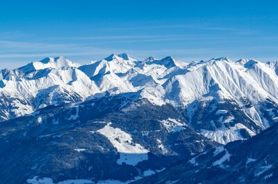 Verschneite Bergkette mit bewaldeten Hängen und Skipisten unter blauem Himmel.