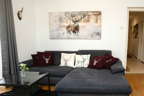 Ein gemütliches Wohnzimmer mit einer grauen Couch und dekorativen Kissen. An der Wand hängt ein Bild von einem Hirsch in naturnaher Umgebung.