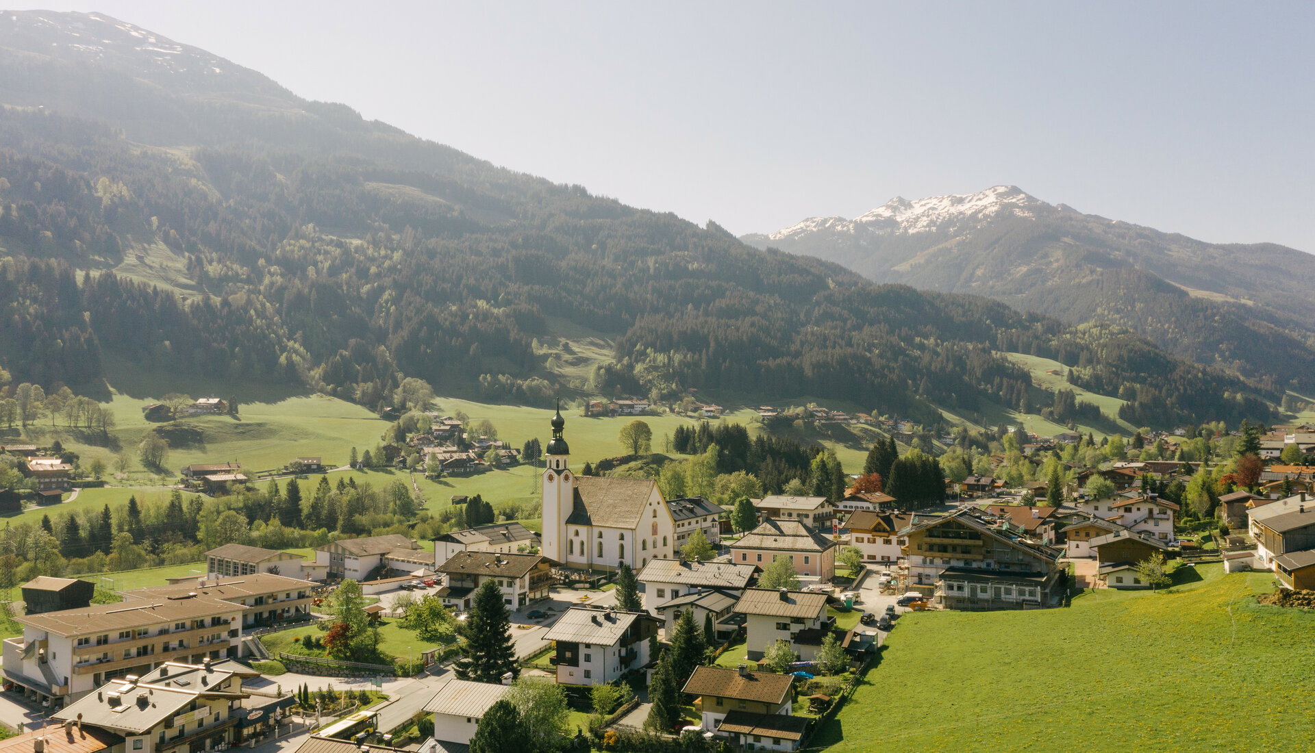 Eine malerische Berglandschaft mit einem kleinen Dorf, umgeben von grünen Wiesen und schneebedeckten Gipfeln im Hintergrund.