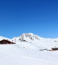 Verschneite Berghütte Hanglalm vor einem felsigen Berggipfel unter blauem Himmel.