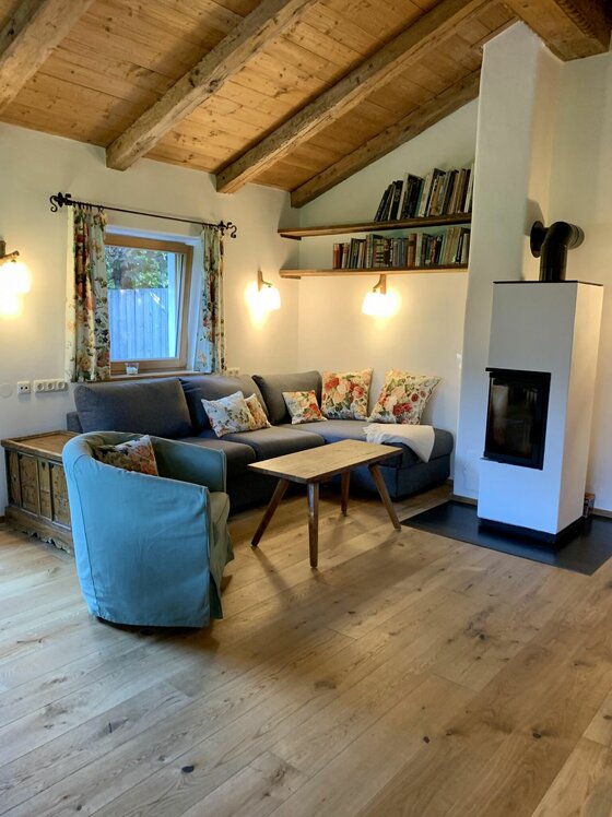 Wohnzimmer mit grauem Ecksofa, Holztisch, Kaminofen und Bücherregalen unter einer Holzbalkendecke.