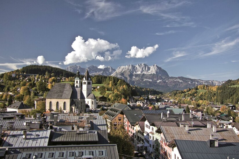 Eine malerische Stadtkulisse mit traditionellen Gebäuden und einer Kirche im Vordergrund. Im Hintergrund erheben sich majestätische Berge unter einem klaren Himmel.