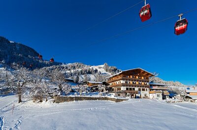 Rote Gondeln einer Seilbahn schweben über einem verschneiten Holzgebäude vor bewaldetem Berghang.