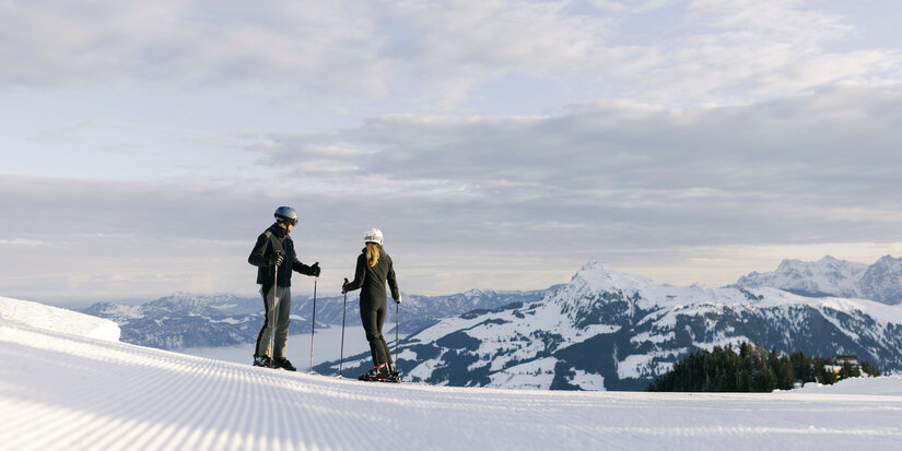 Kitzbühel Ski Resort – Two skiers stand on a snow-covered slope, looking out over the mountains and valley.
