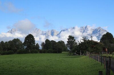 Grüne Wiese mit Bäumen vor einem wolkenverhangenen Gebirgsmassiv unter blauem Himmel.