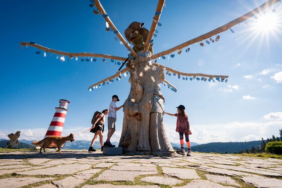 Eine Gruppe von Menschen steht um eine große Statue mit vielen Armen. Im Hintergrund sind Berge und ein Leuchtturm zu sehen, und die Sonne scheint.