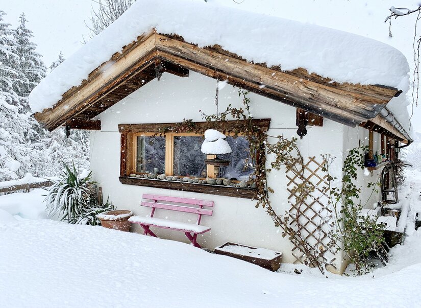 Verschneite Hütte mit rosa Holzbank und schneebedecktem Dach bei leichtem Schneefall.