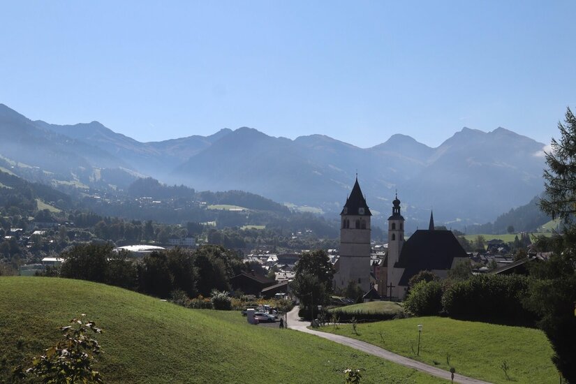 Ortsansicht mit Bergpanorama Blick auf eine Kirche mit zwei Türmen vor einer Gebirgskette unter blauem Himmel.