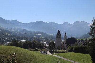 Ortsansicht mit Bergpanorama Blick auf eine Kirche mit zwei Türmen vor einer Gebirgskette unter blauem Himmel.