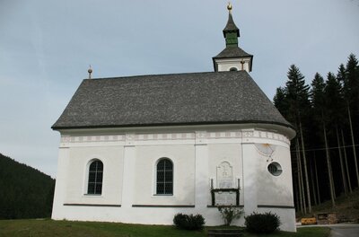 Eine kleine, weiße Kirche mit einem grauen Dach und einem Spitzdach-Glockenturm. Umgeben von Bäumen und einer ruhigen Landschaft.