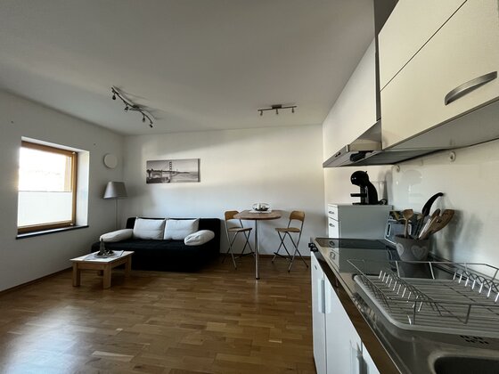 Ein modernes Wohnzimmer mit einem schwarzen Sofa und einem Esstisch für zwei Personen. Die Küche ist hell und gut ausgestattet, mit Holzfußboden und großen Fenstern.