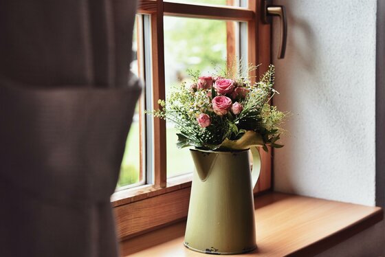 Blumenschmuck am Fenster Blumenstrauß mit rosa Rosen in einer grünen Emaille-Kanne auf einem hölzernen Fensterbrett.