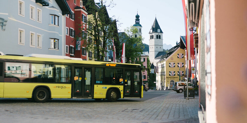 Ein gelber Bus fährt durch die malerische Innenstadt von Kitzbühel, umgeben von bunten Gebäuden und einem klaren Himmel.