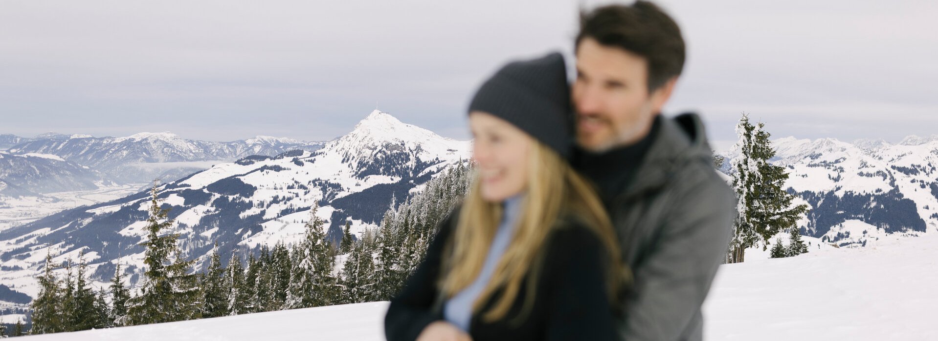 A couple embrace against the backdrop of the snow-covered Kitzbühel Alps