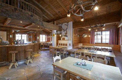 Rustikaler Gastraum mit Holztischen, Bar, Kachelofen und Geweih-Lampen.