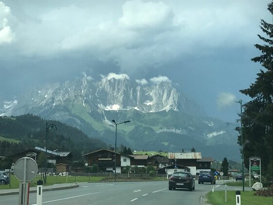 Blick von einer Straße auf eine Ortschaft vor einem massiven, felsigen Gebirgszug unter bewölktem Himmel.