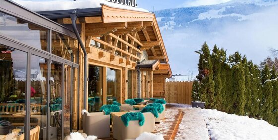 Hofalm Jochberg Außenansicht Außenterrasse einer Holzhütte mit Sesseln und blauen Fellen vor verschneiter Bergkulisse.