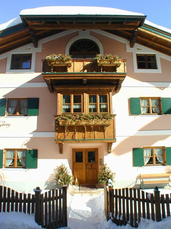 Vorderansicht eines alpinen Hauses mit Holzbalkon, grünen Fensterläden und schneebedecktem Zaun im Vordergrund.