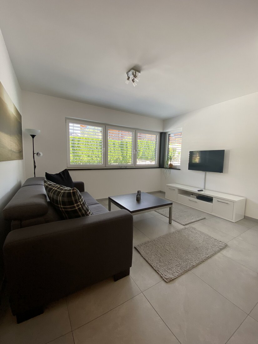 Modernes Wohnzimmer mit dunklem Sofa, Couchtisch, TV an der Wand und hellem Fliesenboden.