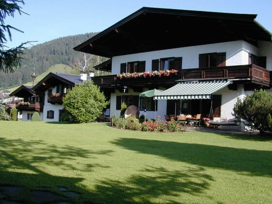Garten des Landhaus Gasteiger Alpines Landhaus mit großem Rasen, Blumenbalkonen und einer Terrasse mit Markise vor bewaldetem Berg.