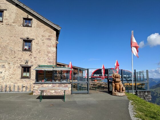 Berghütte aus Stein mit Terrasse, Sonnenschirmen, einer Holzskulptur und einer Seilbahnstation im Hintergrund.