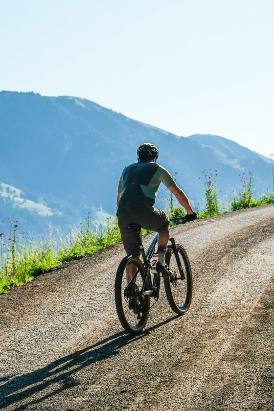 Mountainbiker auf Schotterweg Ein Mountainbiker fährt von hinten gesehen einen steilen Schotterweg bergauf vor einer Bergkulisse.