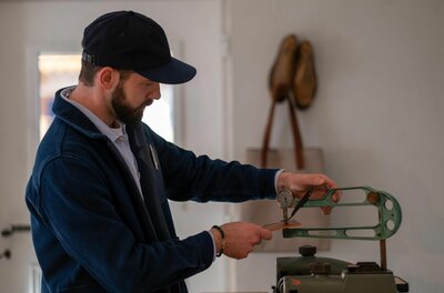 Lederhandwerk im Atelier Ein Mann mit Bart und blauer Kappe misst ein Stück Leder mit einem mechanischen Dickenmesser in einer Werkstatt.