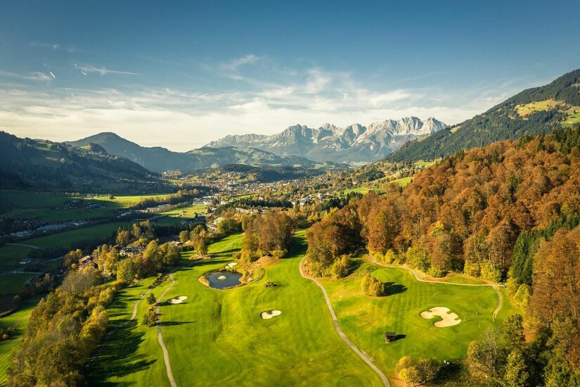 Golfclub Eichenheim Luftaufnahme eines Golfplatzes mit herbstlichen Bäumen vor einem Bergmassiv.