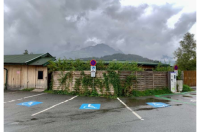 Behindertenparkplatz - Am See 5a Ein Parkplatz mit behindertengerechten Stellplätzen und einem Holzgebäude im Hintergrund. Der Himmel ist bewölkt, und im Hintergrund sind Berge sichtbar.