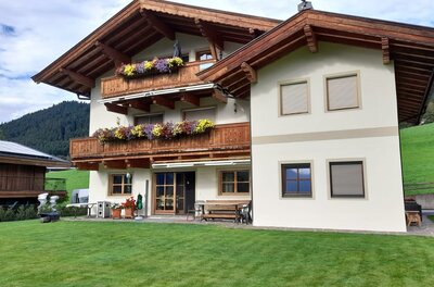 1000095352 Ein traditionelles Alpenhaus mit Holzbalustraden und blühenden Blumenkästen. Im Hintergrund liegen grüne Wiesen und bewaldete Berge.