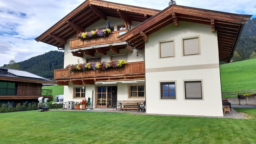 1000095352 Ein traditionelles Alpenhaus mit Holzbalustraden und blühenden Blumenkästen. Im Hintergrund liegen grüne Wiesen und bewaldete Berge.