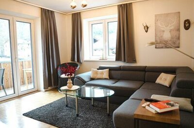 605345669 Ein modernes Wohnzimmer mit einer grauen Couch und einem Glastisch. Die großen Fenster lassen viel Licht herein und zeigen eine gemütliche Atmosphäre.