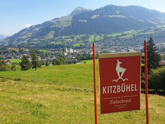 Rote Hinweistafel Zielschuss Kitzbühel auf einer grünen Wiese mit Blick auf den Ort und Berge.