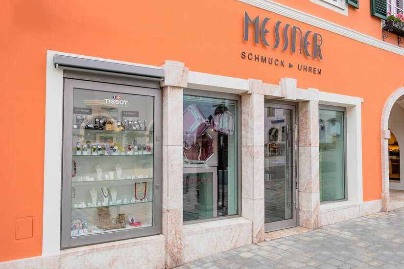 Messner Schmuck und Uhren Außenansicht eines Schmuck- und Uhrengeschäfts mit orangefarbener Fassade und Schaufensterauslagen.