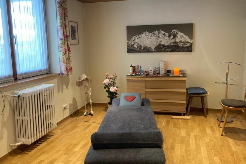 Praxisraum Monika Ober Ein Behandlungsraum mit Massageliege, Holzkommode und einem Gebirgsbild an der Wand.