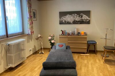 Praxisraum Monika Ober Ein Behandlungsraum mit Massageliege, Holzkommode und einem Gebirgsbild an der Wand.