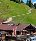 Berghütte aus Holz mit Sonnenterrasse vor einem grünen Grashang mit Schotterweg.